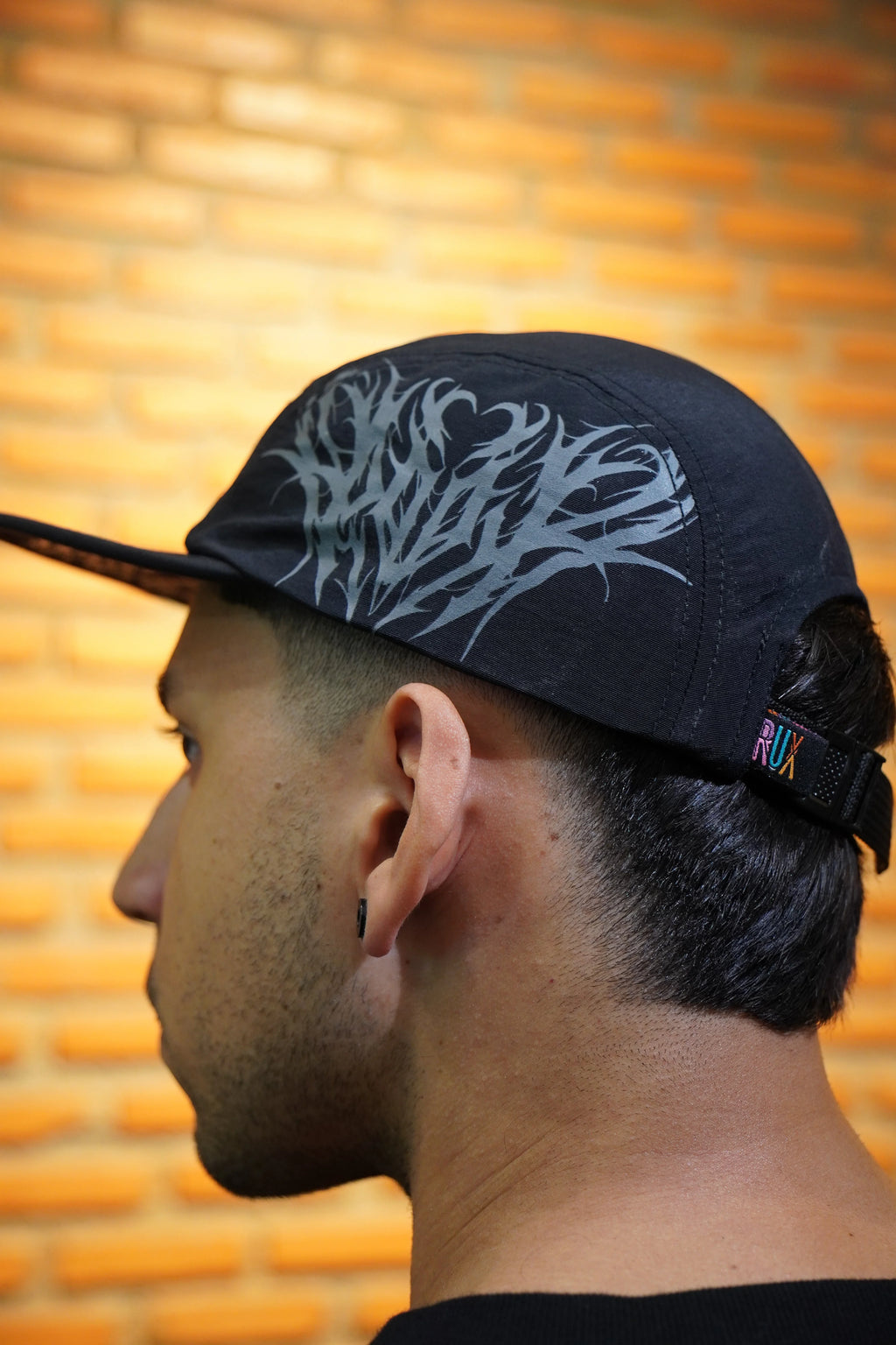 Model wearing a black five panel cap with gothic print and adjustable strap