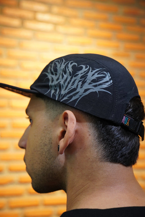 Model wearing a black five panel cap with gothic print and adjustable strap