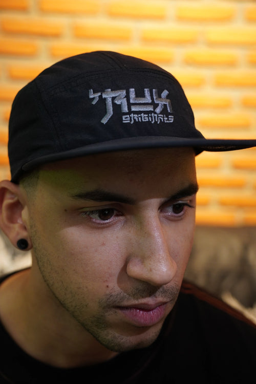Man wearing a black five panel cap with white embroidered text, brick wall background.