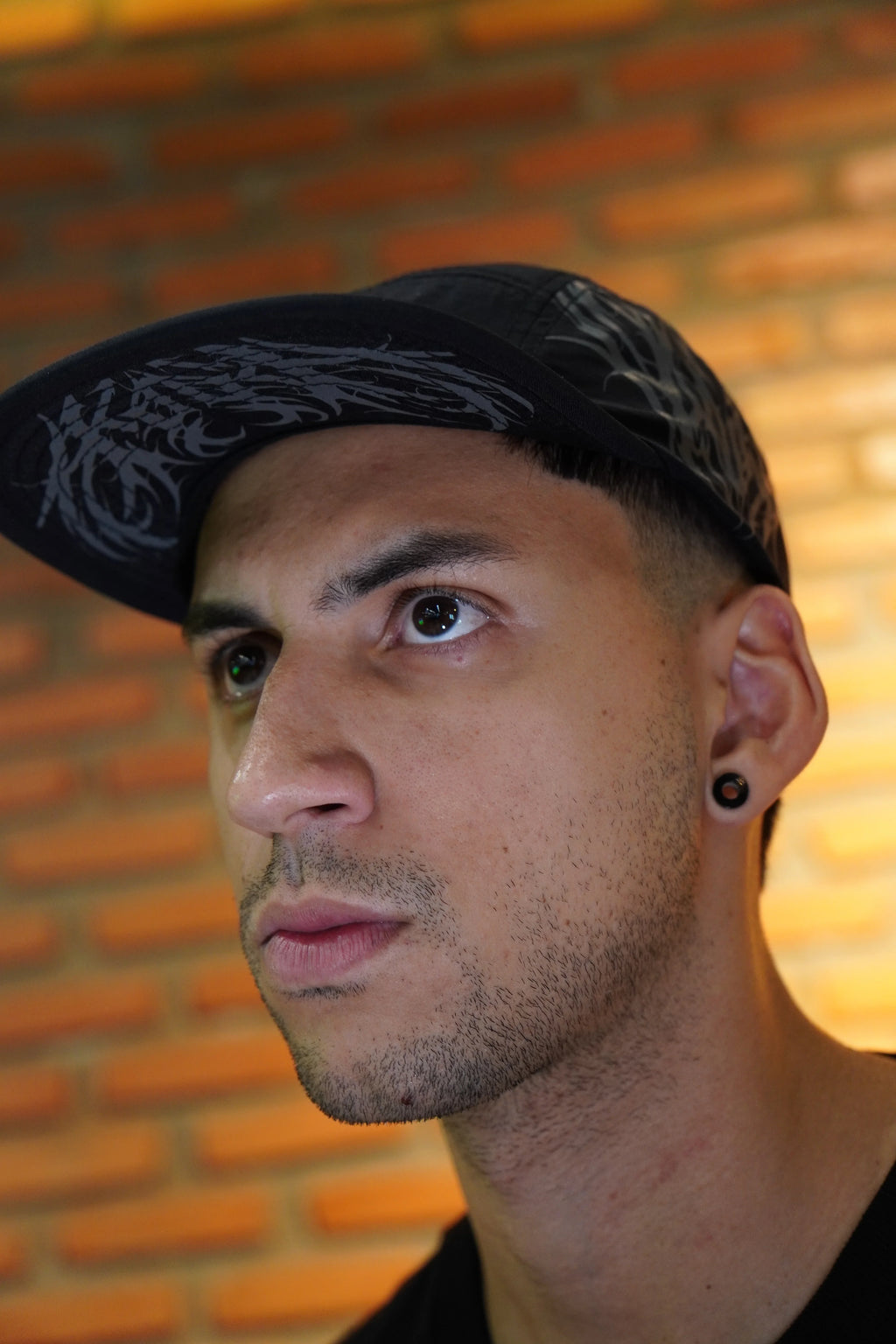 Model wearing a black five panel cap with graphic design, brick wall background