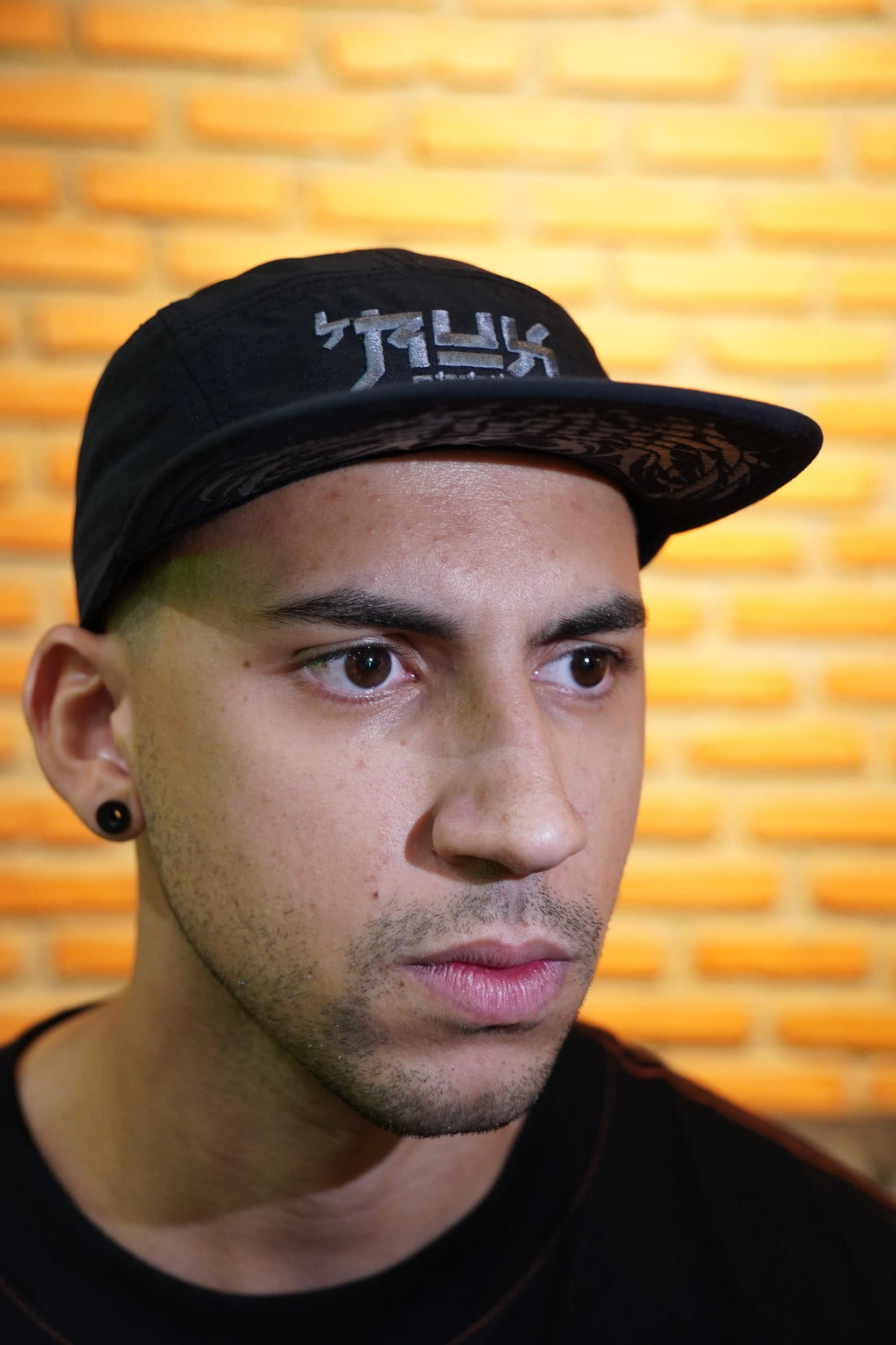 Close-up of model wearing a black five-panel hat with gothic lettering, brick wall background