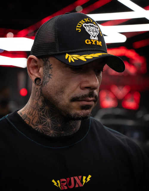 Model wearing a tattooed neck, black mesh cap, and printed t-shirt in a gym setting