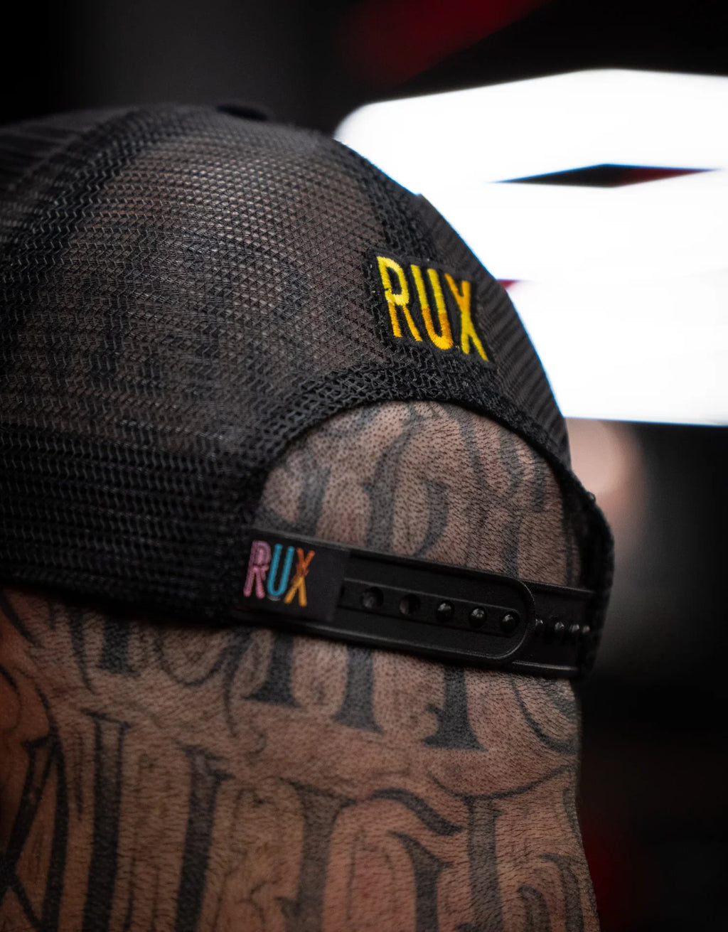 Close-up of model wearing black mesh trucker hat with RUX logo, tattooed head visible