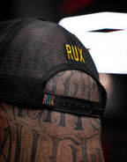 Close-up of model wearing black mesh trucker hat with RUX logo, tattooed head visible