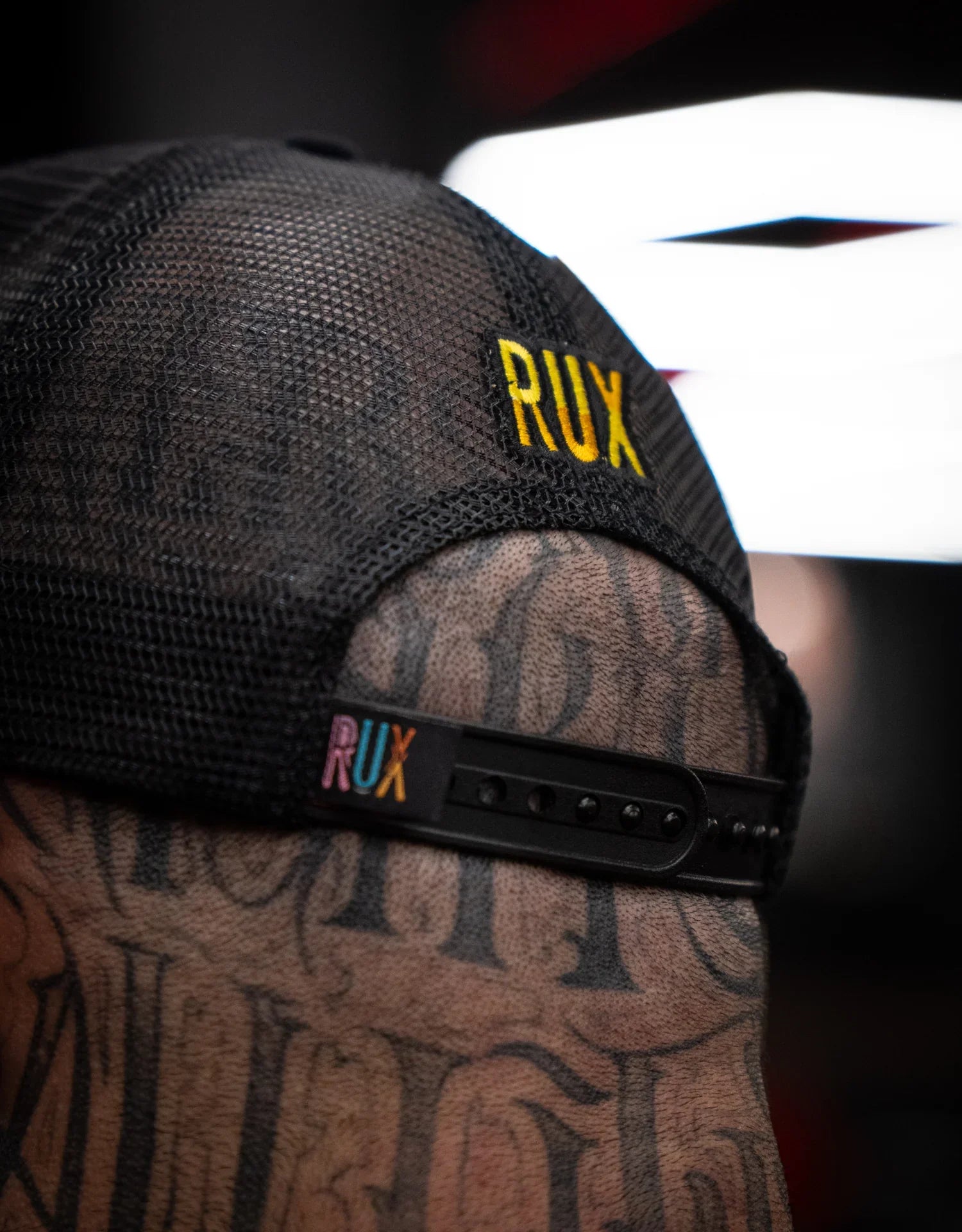 Close-up of model wearing black mesh trucker hat with RUX logo, tattooed head visible
