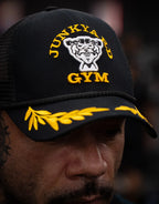 Person wearing a black trucker cap with yellow Junkyard Gym embroidery and laurel detail