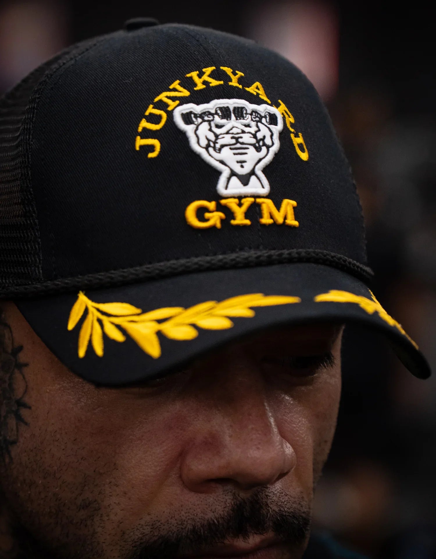 Person wearing a black trucker cap with yellow Junkyard Gym embroidery and laurel detail