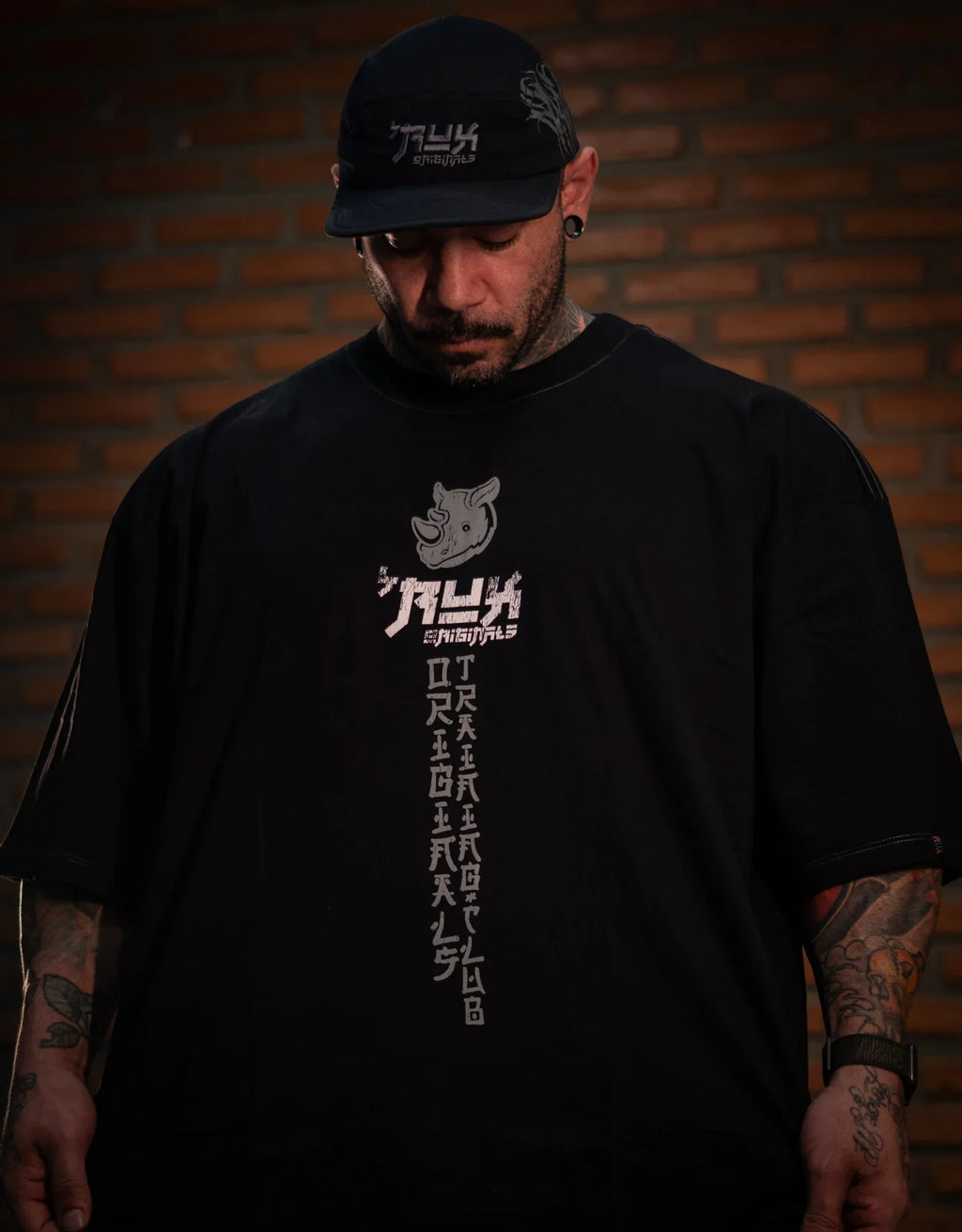 Model wearing black oversized t-shirt with graphic print and black cap, brick wall background