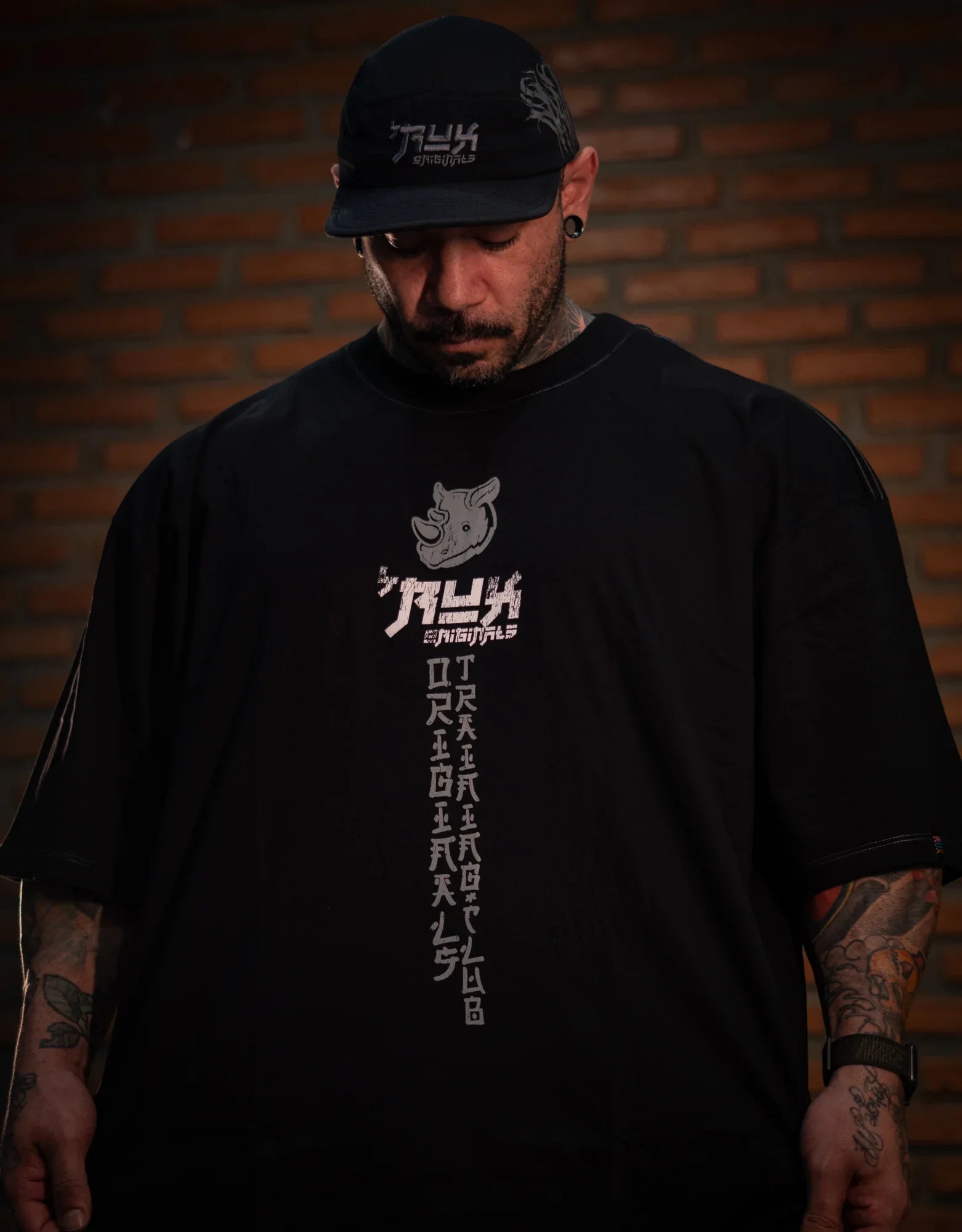 Model wearing black oversized t-shirt with graphic print and black cap, brick wall background