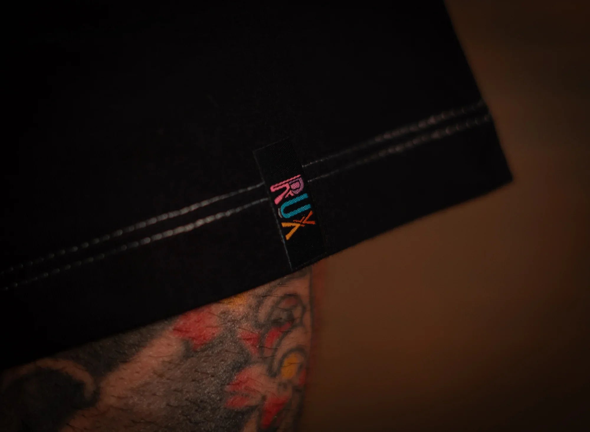 Close-up of model wearing black pants with colorful logo tag and tattooed leg visible