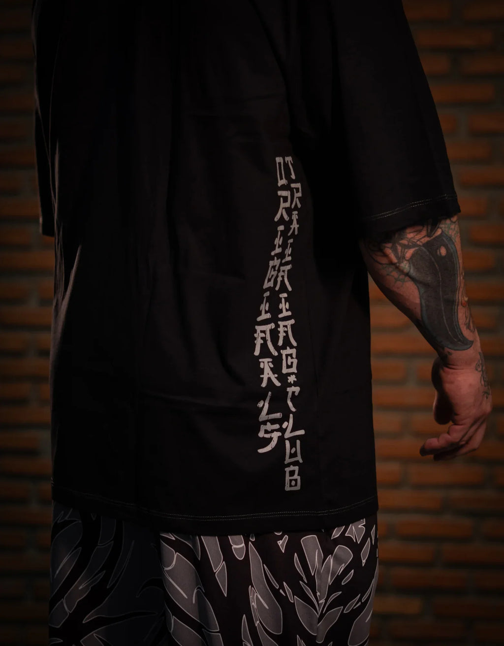 Model wearing patterned black and gray pants, side tattoo visible, brick wall background