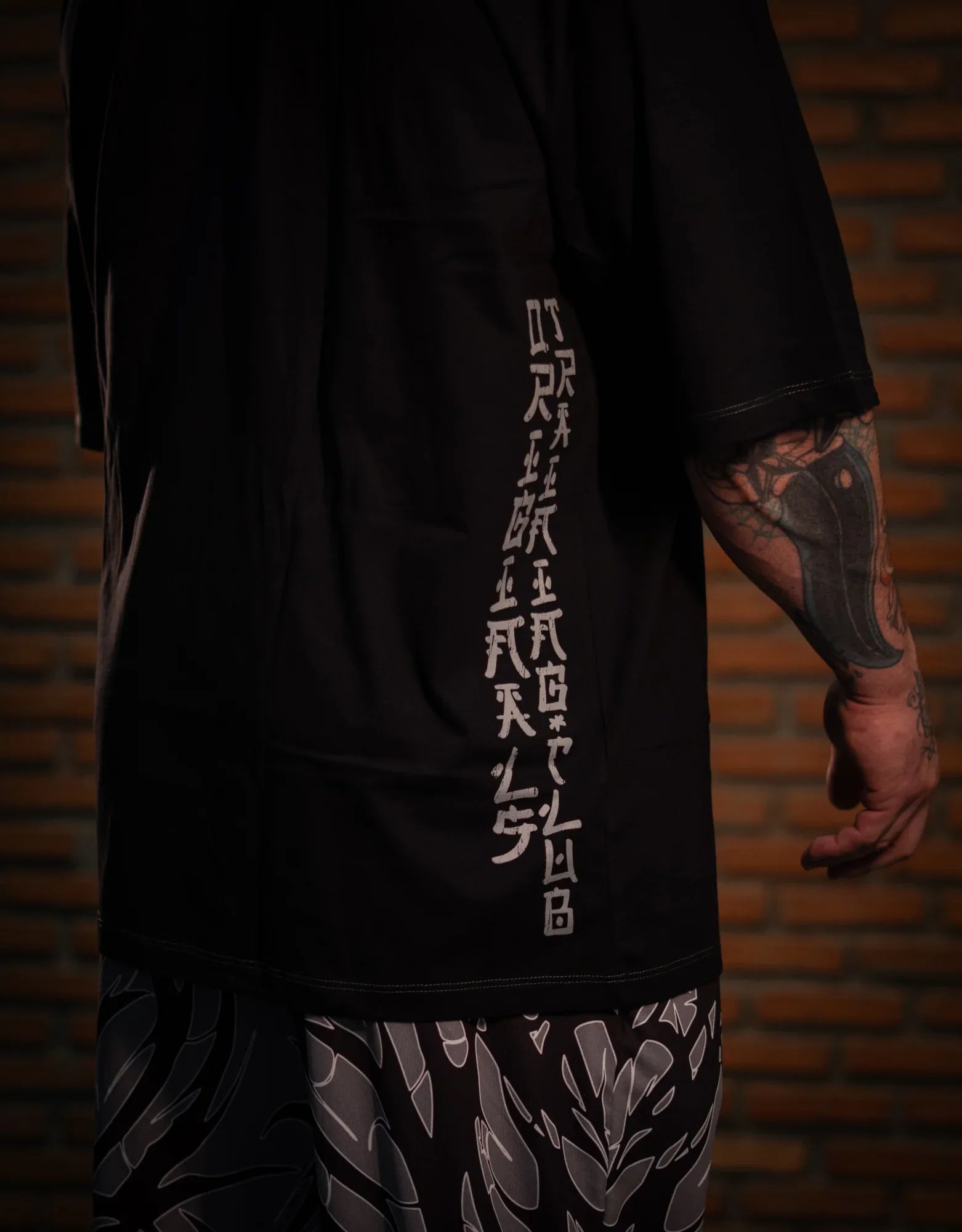 Model wearing patterned black and gray pants, side tattoo visible, brick wall background