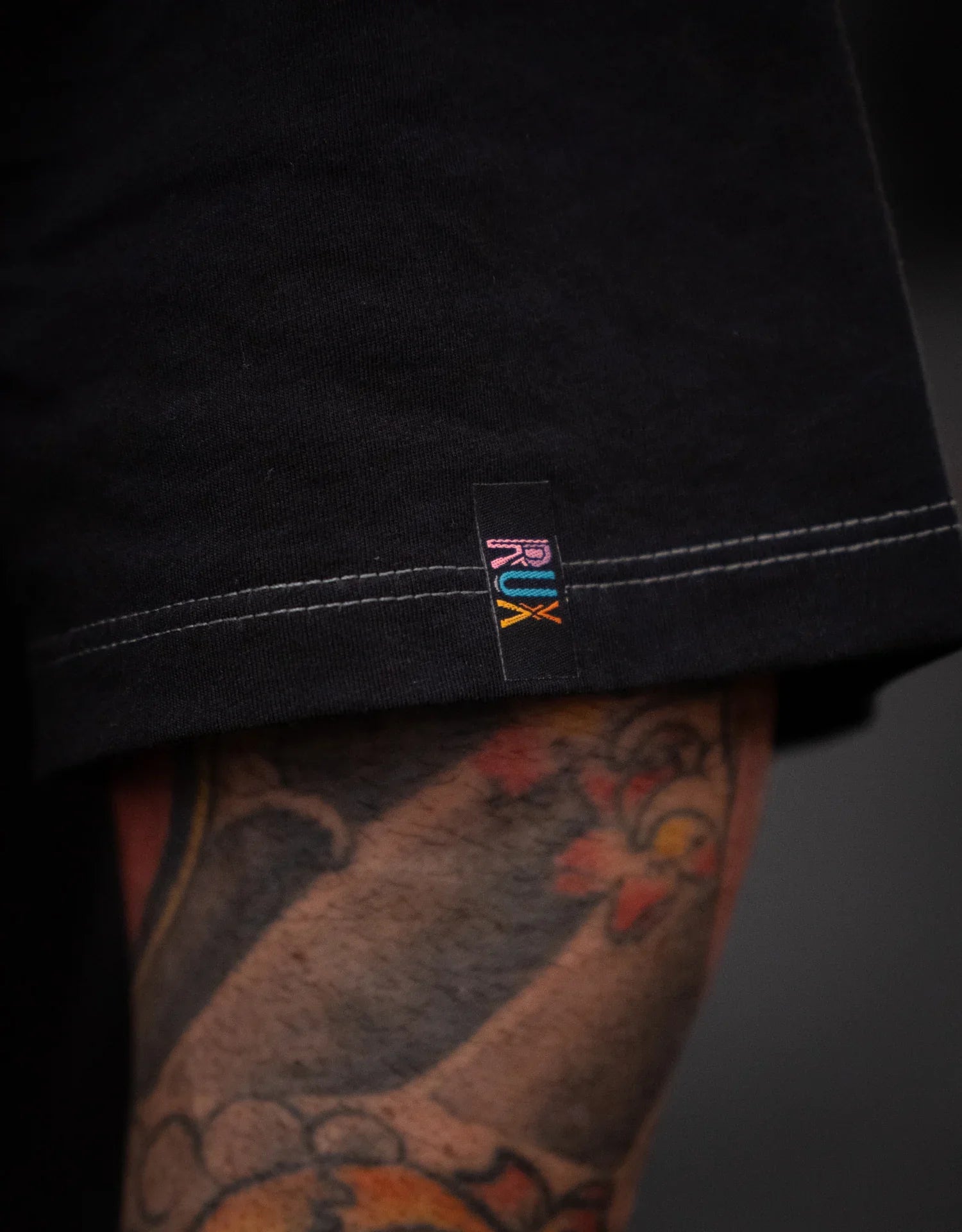 Model wearing black pants, visible hem with stitched label, tattooed leg showing