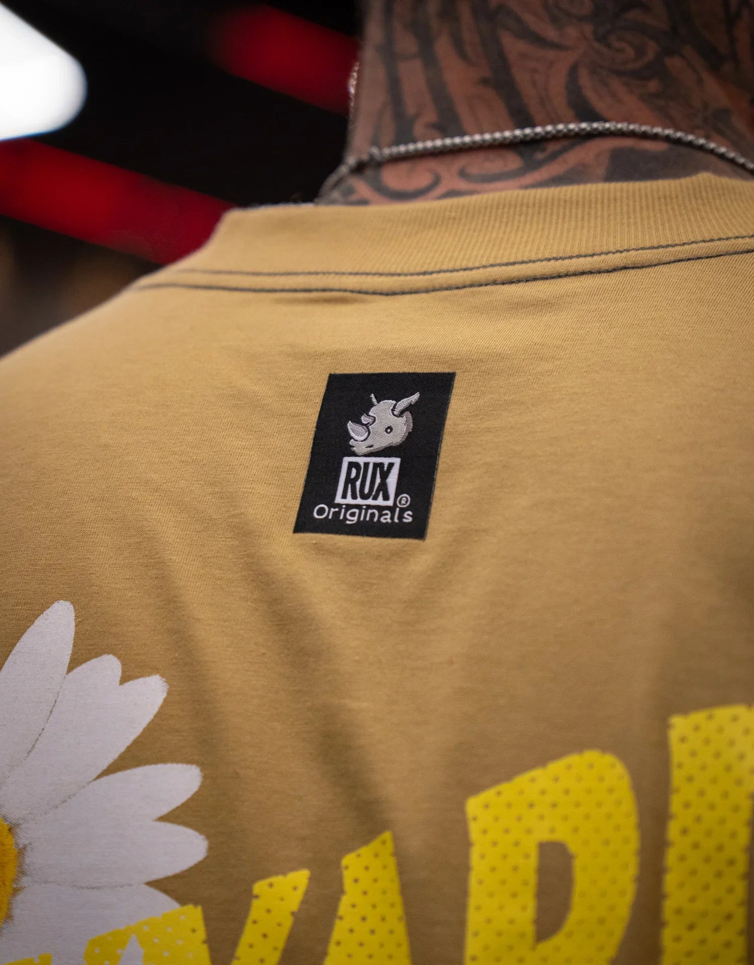 Close-up of person wearing tan graphic t-shirt with visible Rux Originals tag and tattooed neck