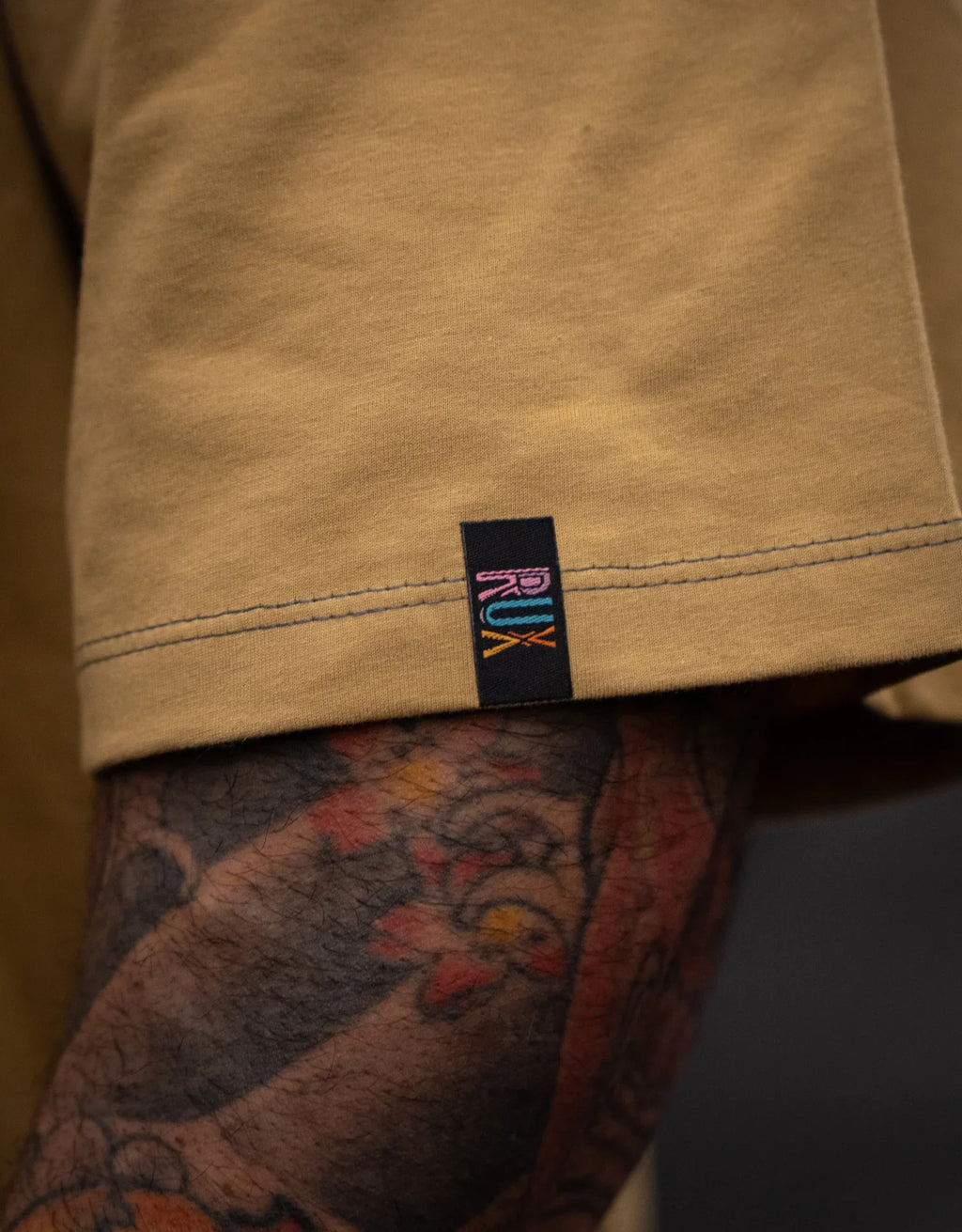 Model wearing tan pants with visible hem and small black tag, leg with colorful tattoos