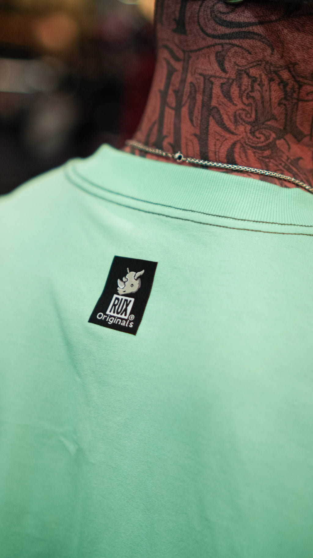 Model wearing turquoise shirt with visible neck tattoos and a logo tag on the back